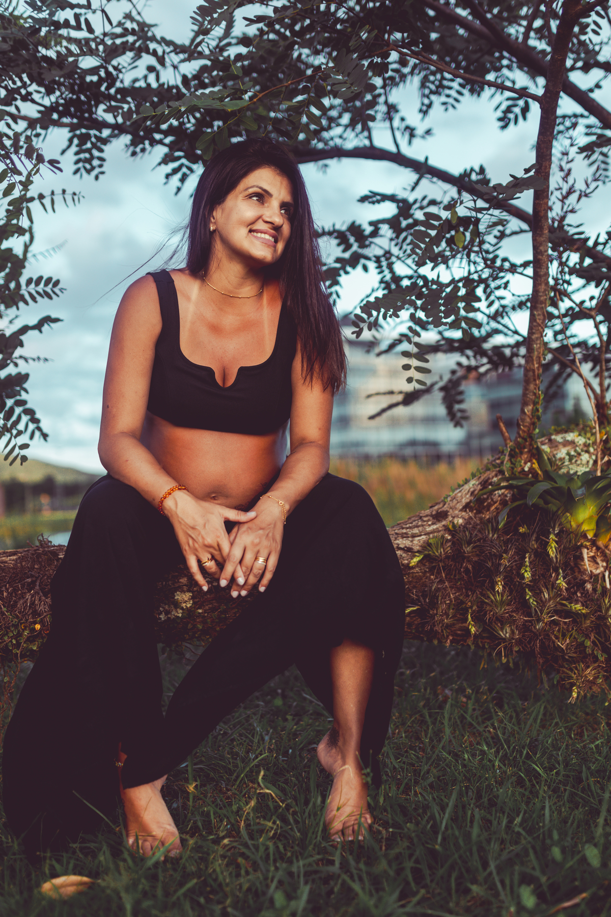 gestante de top e calça preta sentada em galho de árvore olhando pro lado e sorrindo