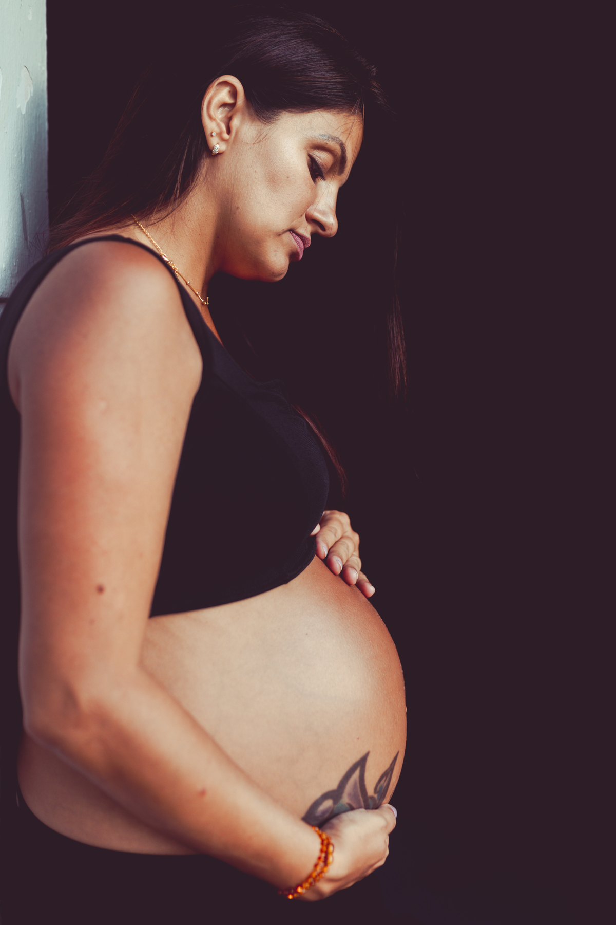 mulher gestante encostada de lado em parede com fundo preto segurando a barriga