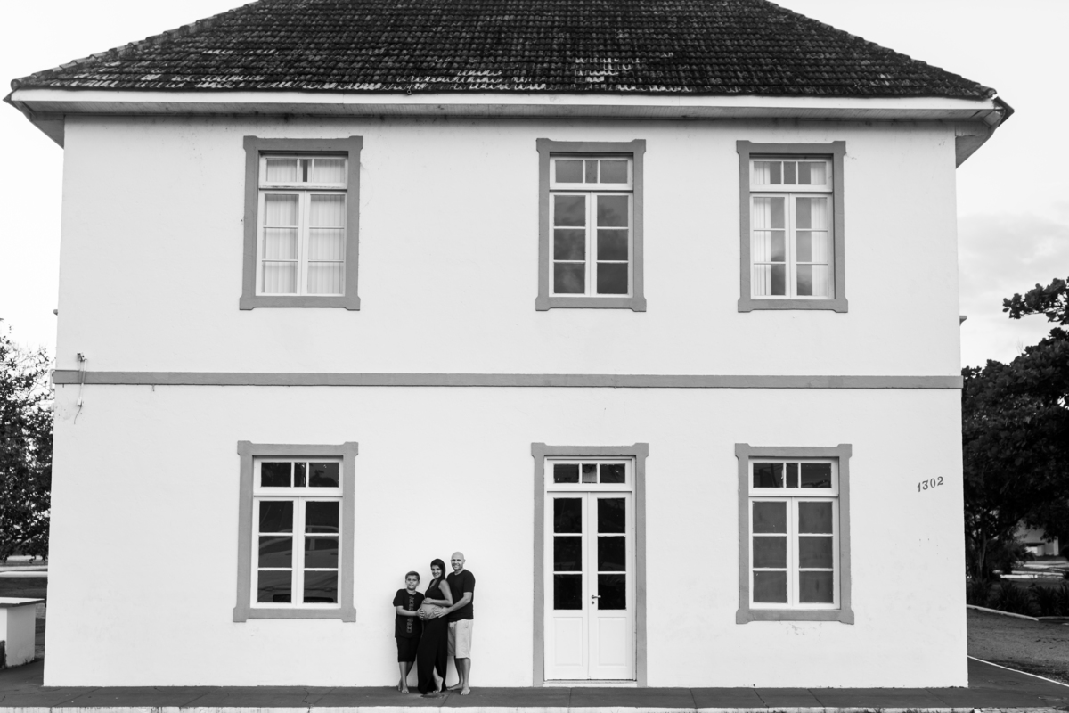 foto em preto e branco de gestante, marido e filho em frete a casa de 2 andares