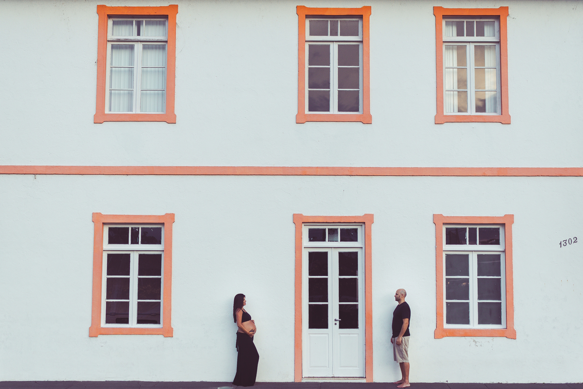 foto de esposa gestante e marido um de frente para o outro, em frente a casa branca com detalhes em laranja