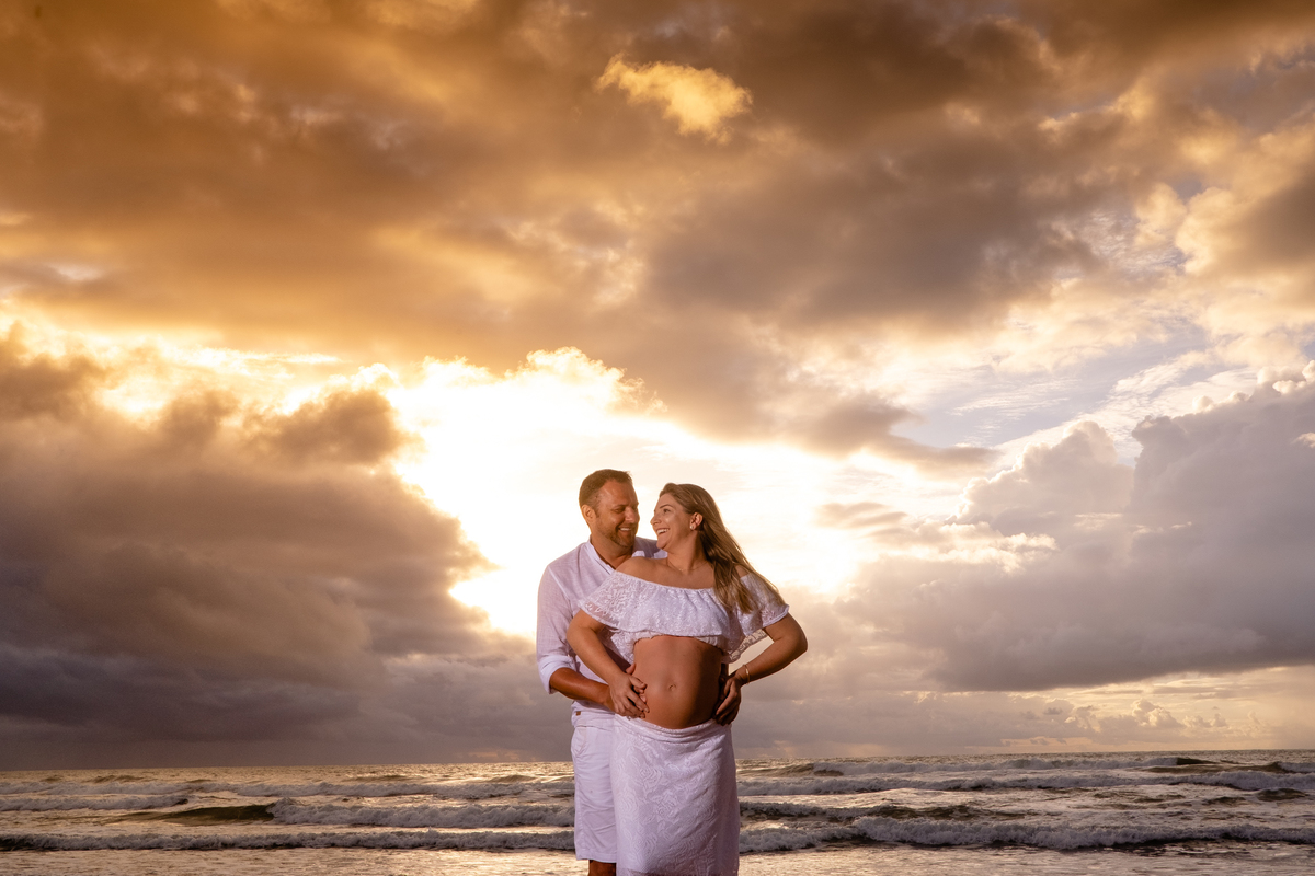 Ensaio de gestante com marido sorrindo na praia e nascer do sol.