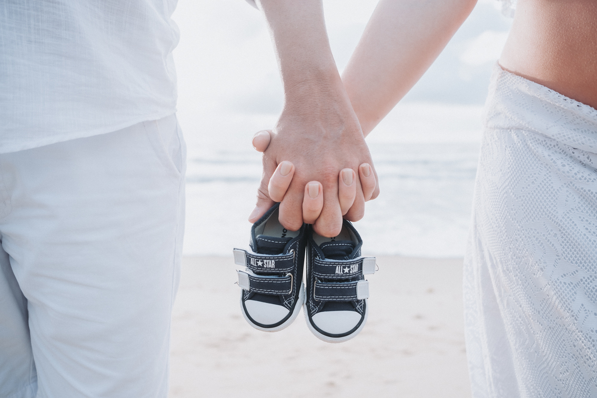 Detalhes das mãos segurando sapatinho do bebê no ensaio de gestante na praia e no nascer do sol