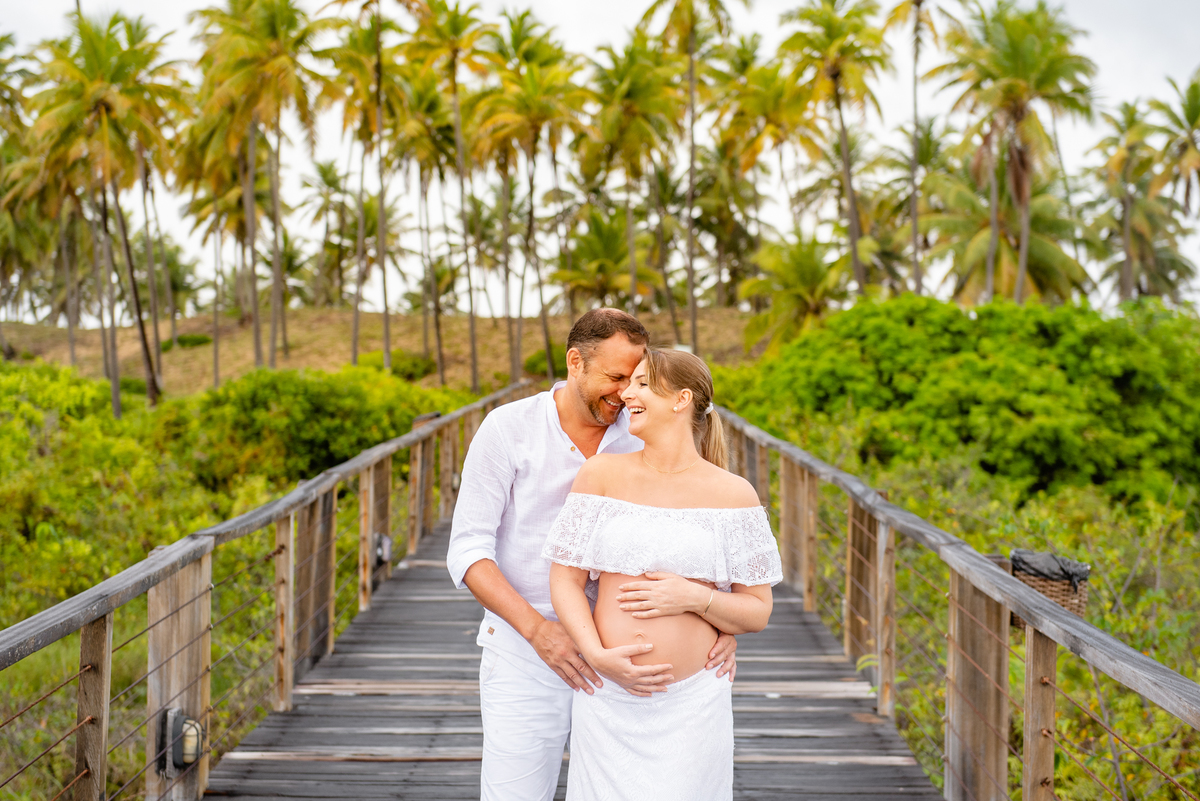 foto no ensaio de gestante na praia com coqueiros e casal se abraçando
