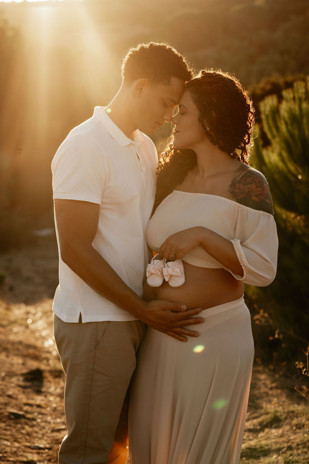 Casal grávido abraçado com as mãos sobre a barriga durante uma sessão de maternidade ao pôr do sol.