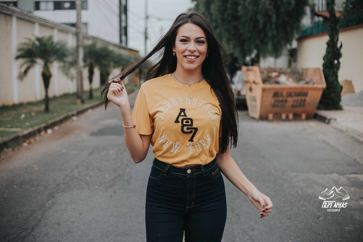 Livia Melo caminhando pela rua durante um ensaio fotografico em Betim em que ela esta mexendo no cabelo e sorrindo