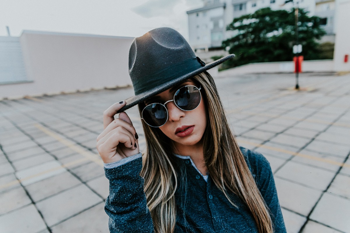 Ensaio feminino no qual a modelo está usando oculos escuros e chapéu fedora em um ensaio no estácionamento