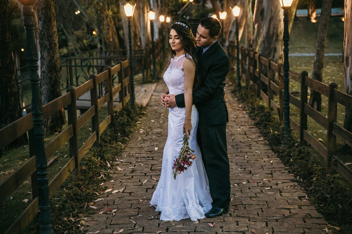 Retrato criativo de pós casamento rustico no pós do sol no qual o noivo está abraçando a noiva por trás cercado de arvores 