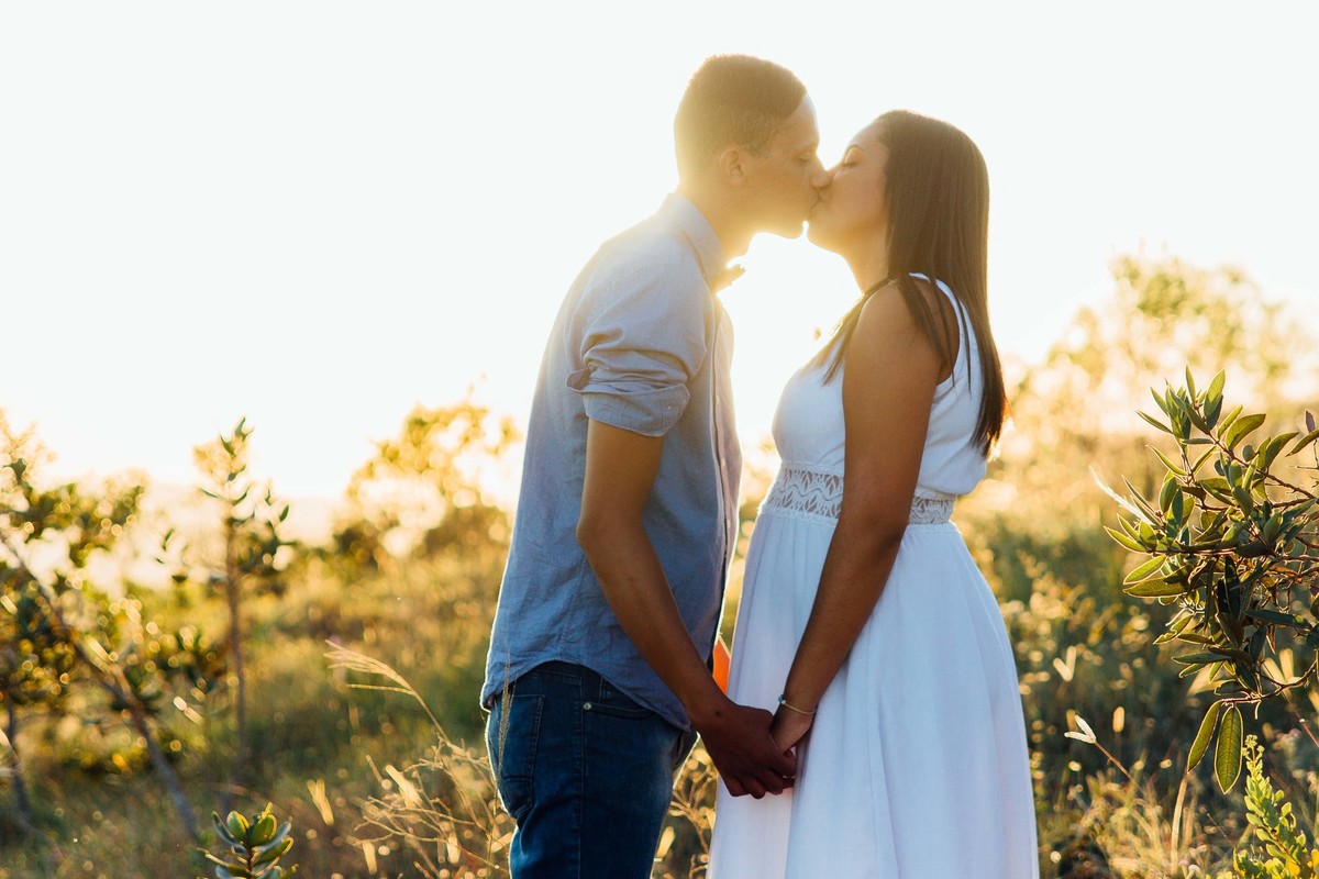 Noivos se beijando no por do sol de mão dadas, a noiva usa vestido branco e o noivo uma camisa azul, eles fazem pose para as fotos