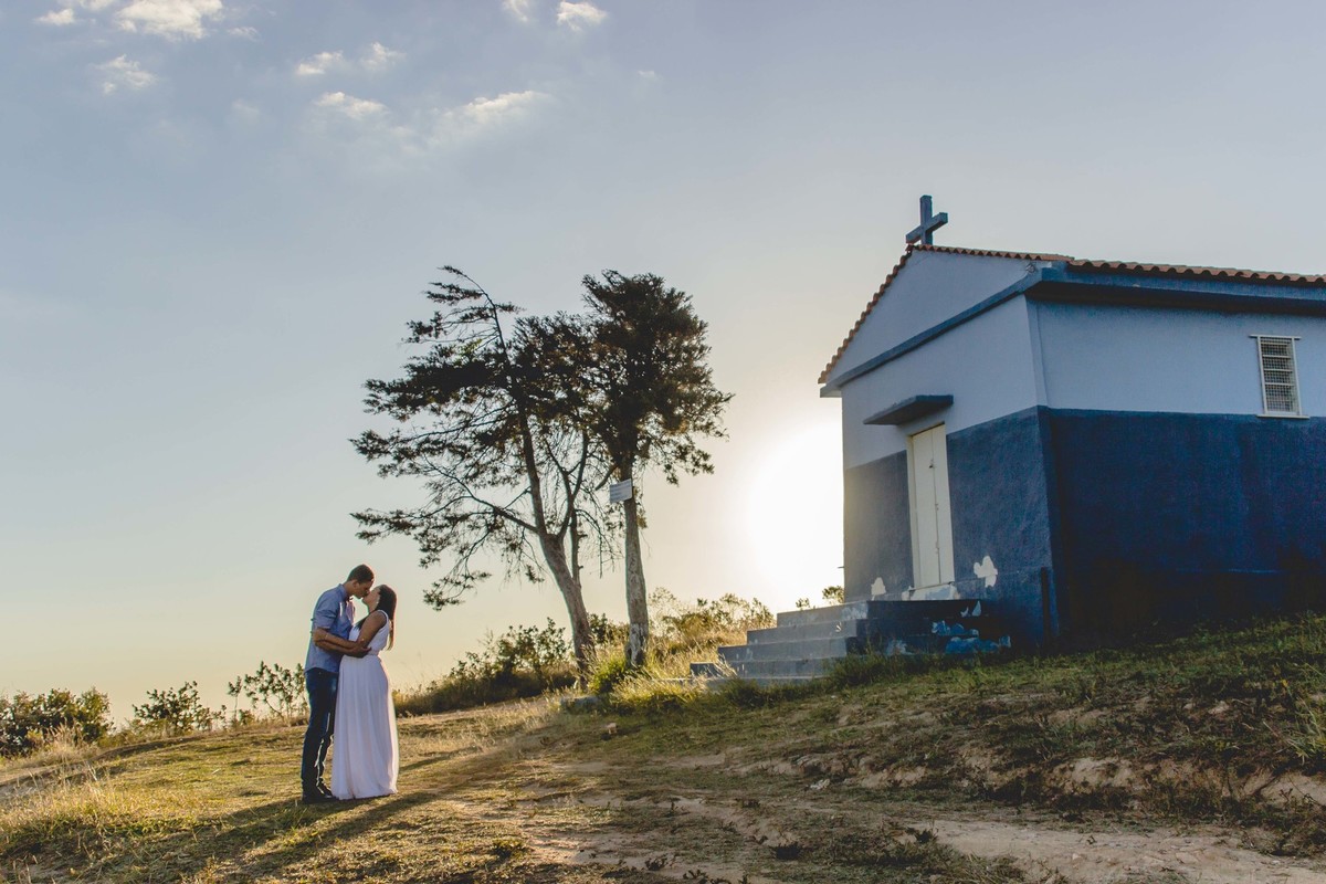 fotos no por do sol de recem casados, na frente de uma igreja azul, noiva com vestido branco e o noivo com blusa social e gravata borboleta, fotos na frente da capela