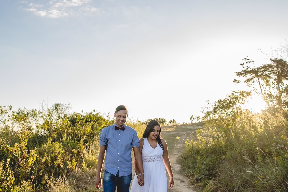 fotos no por do sol de recem casados, na frente de uma igreja azul, noiva com vestido branco e o noivo com blusa social e gravata borbleta, caminhando por uma estrada no alto da montanha