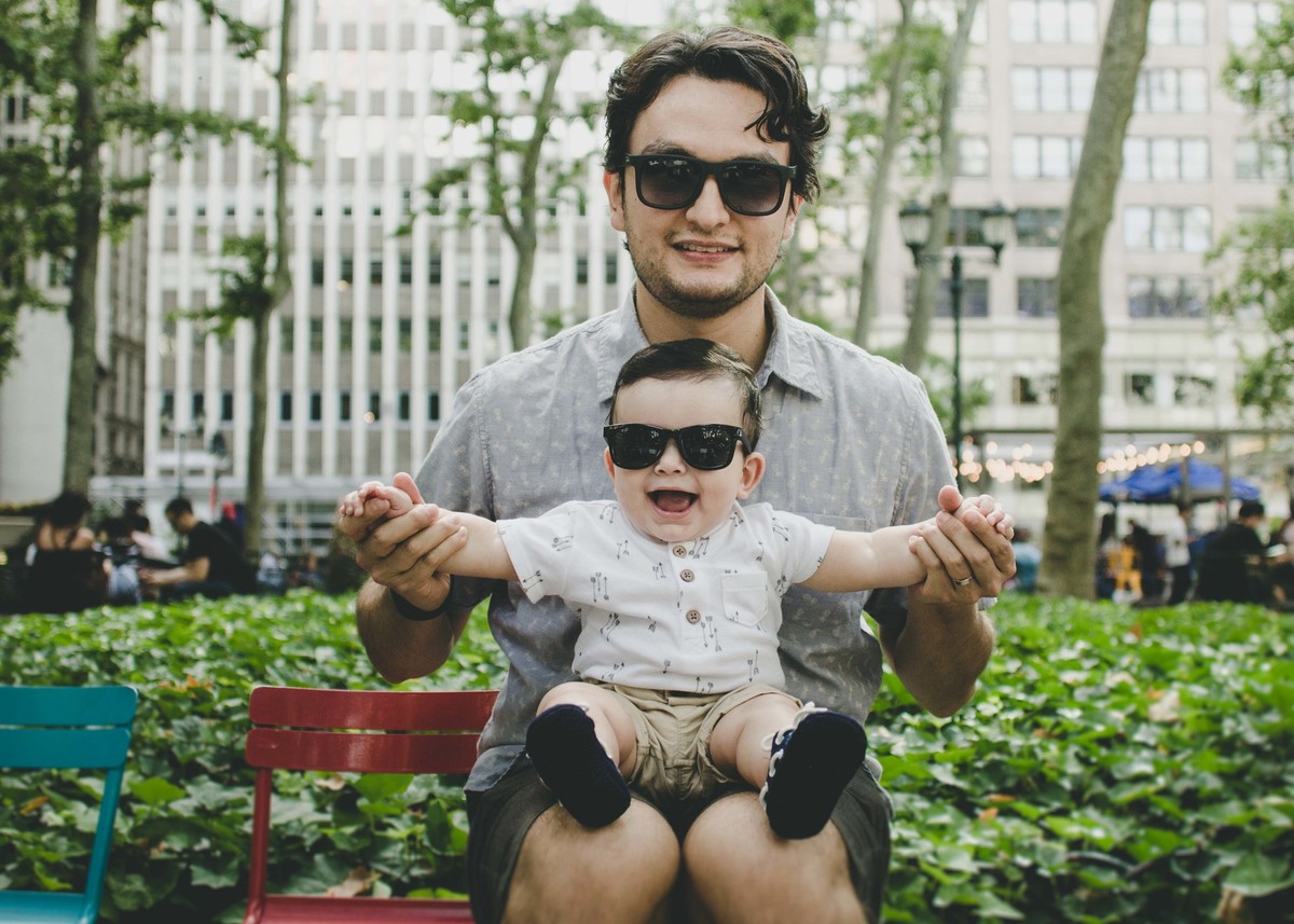 Ensaio fotográfico de família em Nova York, pai e filho com óculos escuro sorrindo, pai segurando filho no colo levantando os braços do filho em um parque