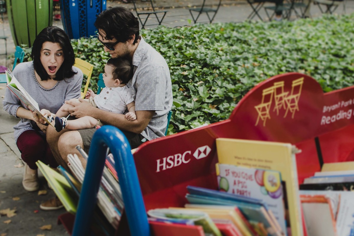 Ensaio fotográfico de família em Nova York, pai segurando filho no colo, mãe lendo livro para o filho, estante de livros infantis, em uma praça ou parque