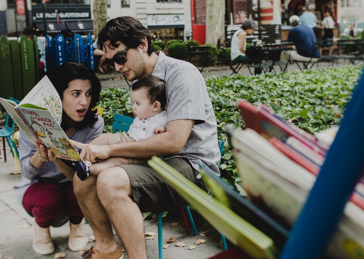 Ensaio fotográfico de família em Nova York, pai segurando filho no colo, mãe lendo livro para o filho, estante de livros infantis, em uma praça ou parque