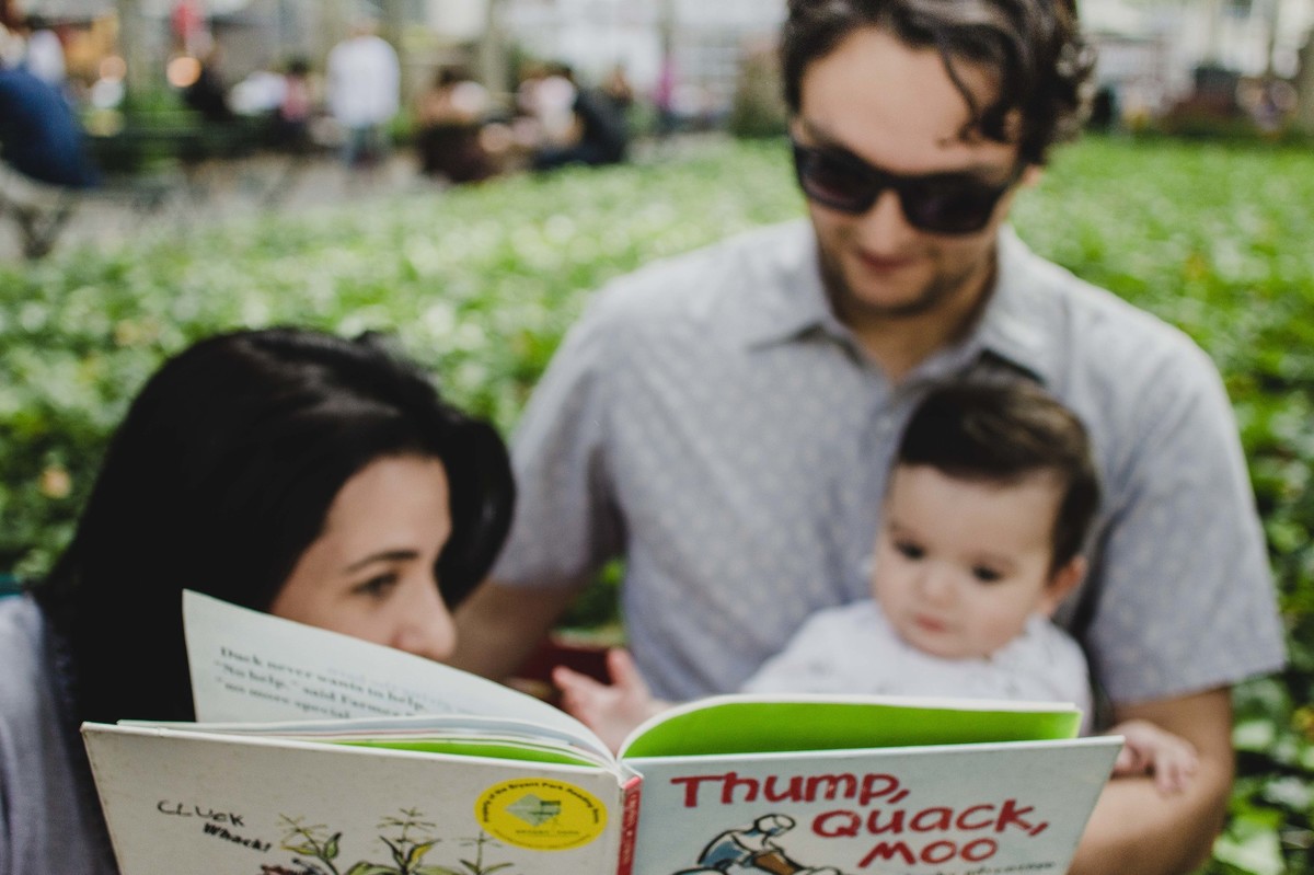 Ensaio fotográfico de família em Nova York, pai segurando filho no colo, mãe lendo livro para o filho  em uma praça ou parque