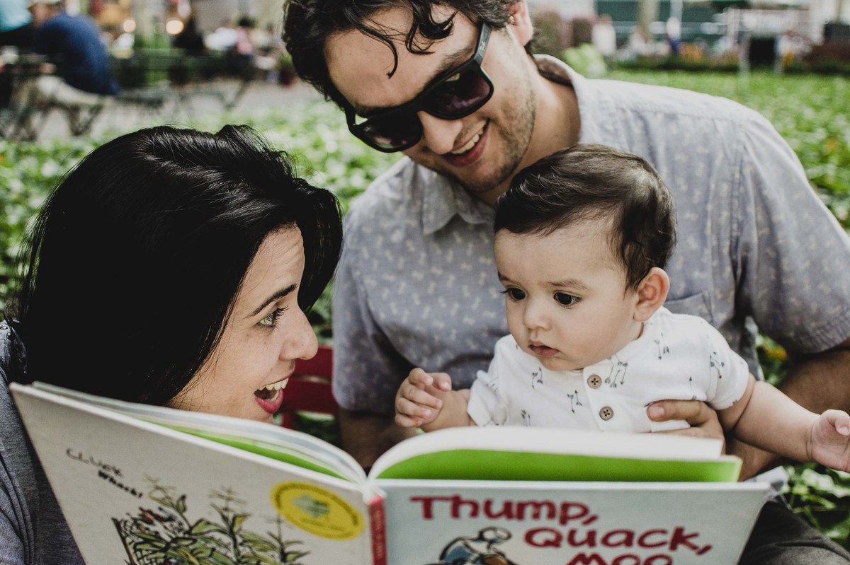 Ensaio fotográfico de família em Nova York, pai segurando filho no colo, mãe lendo livro para o filho  em uma praça ou parque, mãe sorri para o filho