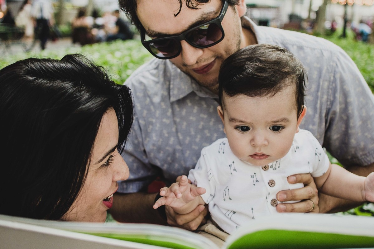 Ensaio fotográfico de família em Nova York, pai segurando filho no colo, mãe lendo livro para o filho  em uma praça ou parque, mãe sorri para o filho, filho olha para o livro espantado