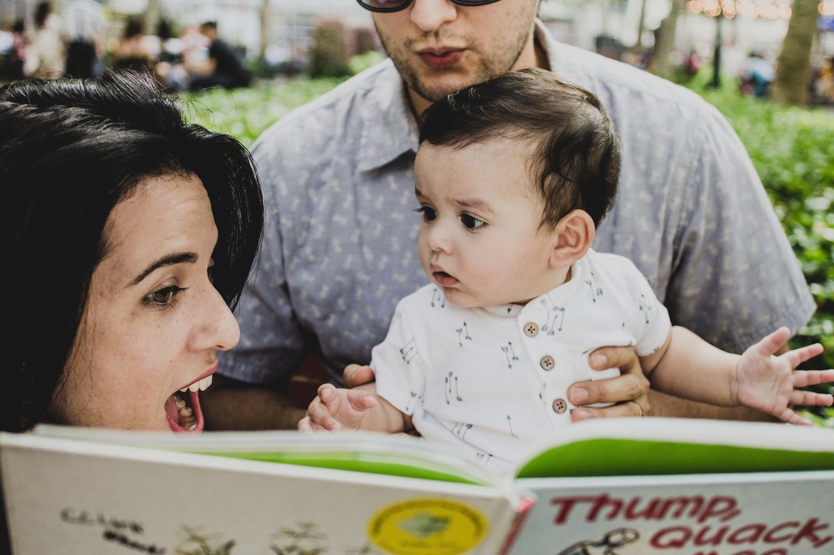 Ensaio fotográfico de família em Nova York, pai segurando filho no colo, mãe lendo livro para o filho  em uma praça ou parque, mãe sorri para o filho, filho olha para mãe espantado