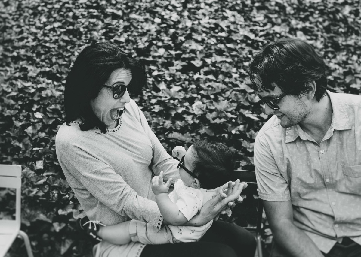Ensaio fotográfico de família em Nova York, mãe segurando filho no colo, mãe sorri, filho sorri para mãe, pai observa, eles estão em parque, foto preto e branco