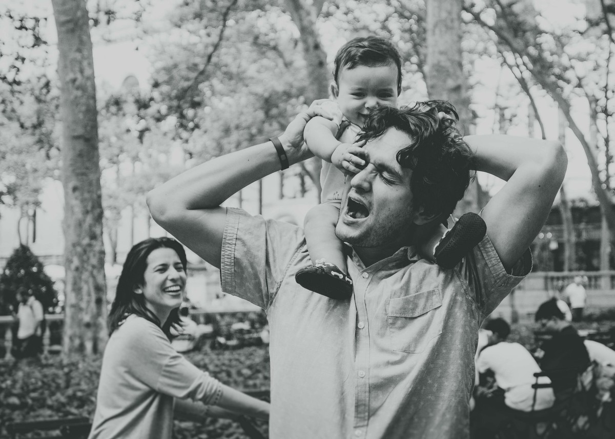 Ensaio fotográfico de família em Nova York, pai segura filho nos ombros, filho coloca mãos no olho do pai, mãe observa e sorri, foto preto e branco
