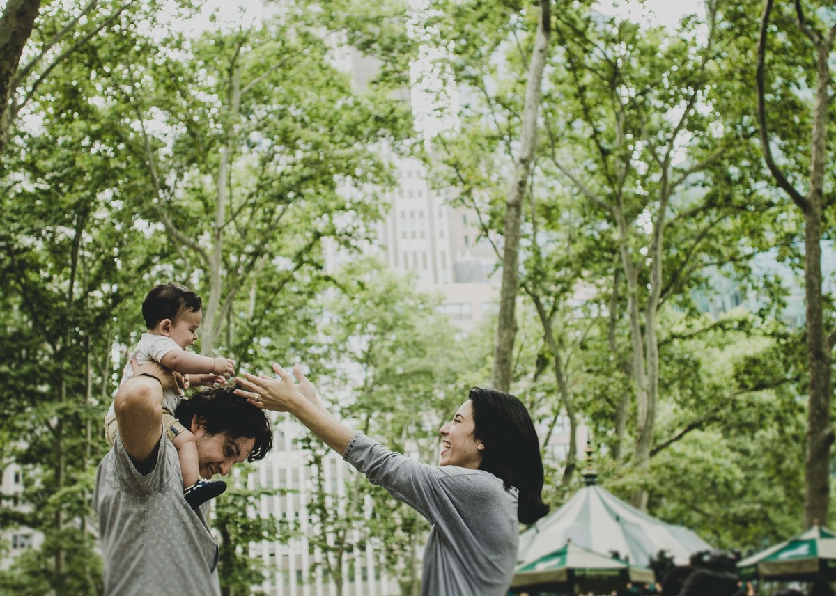 Ensaio fotográfico de família em Nova York, pai segura filho nos ombros, filho sorri, mãe estende os braços chamando o filho