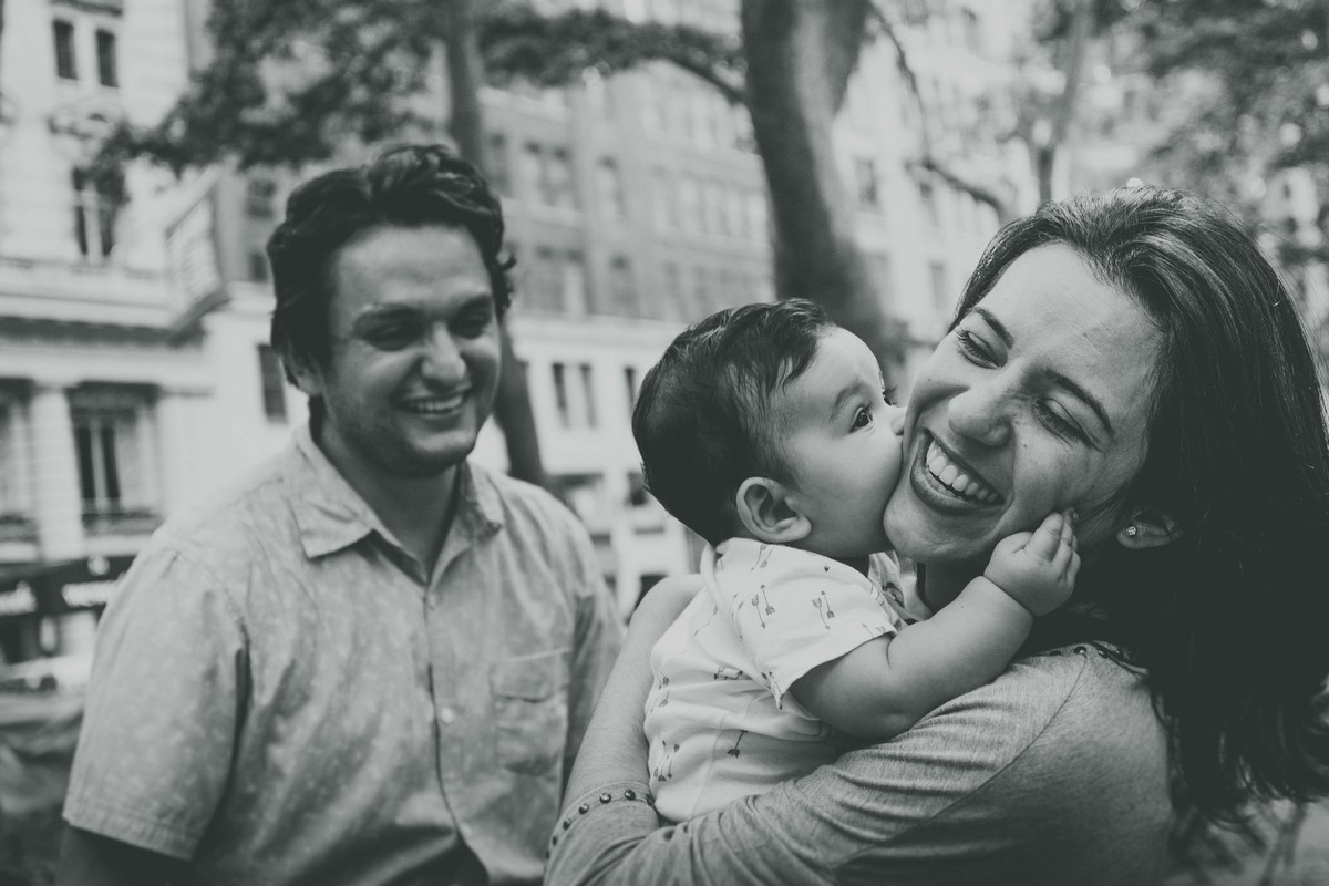 Ensaio fotográfico de família em Nova York, mãe segura filho no colo, filho morde a bochecha da mãe, pai observa e sorri, praça ou parque, foto preto e branco