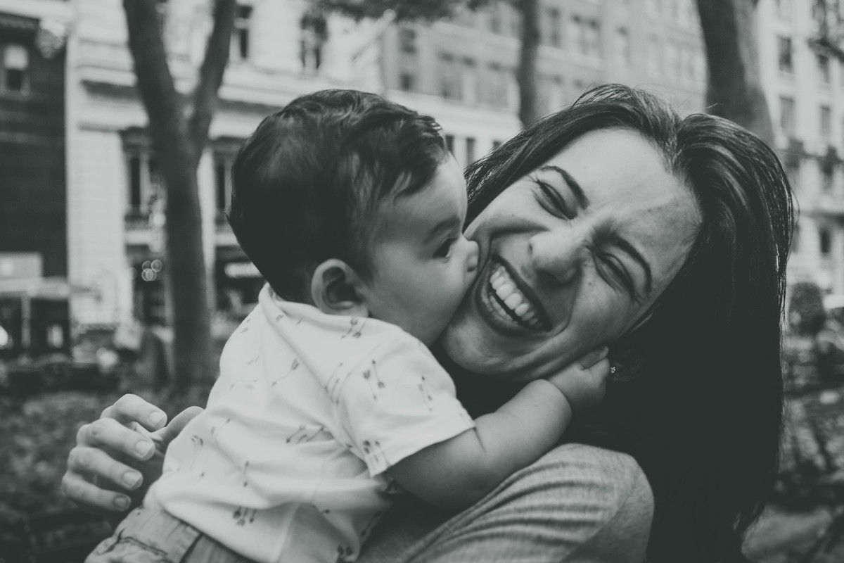 Ensaio fotográfico de família em Nova York, mãe segura filho no colo, filho morde a bochecha da mãe, praça ou parque, foto preto e branco