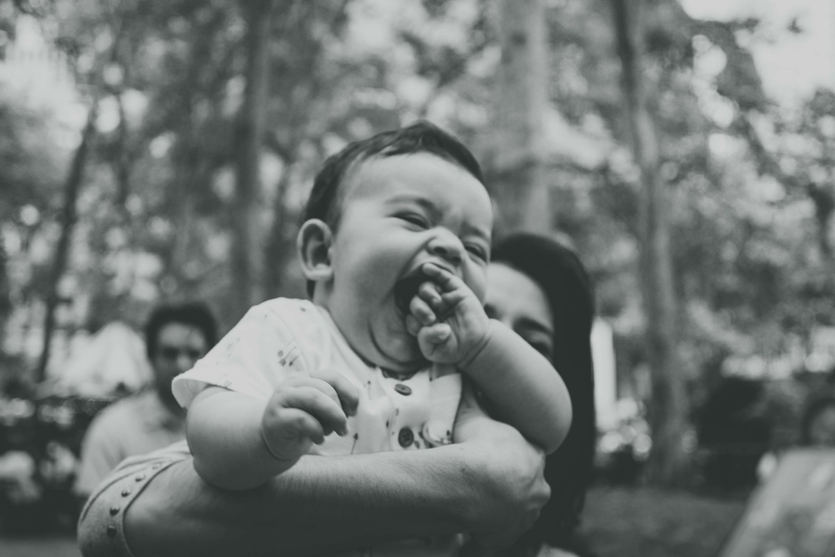 Ensaio fotográfico de família em Nova York, mãe segura filho no colo, filho sorri e morde a própira mão, praça ou parque, foto preto e branco