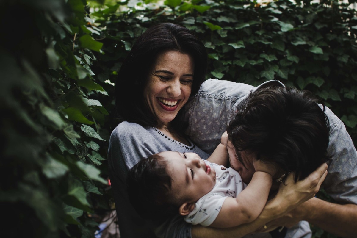 Ensaio fotográfico de família em Nova York, mãe segura filho no colo, pai faz cócegas em filho que sorri, mãe observa e sorri