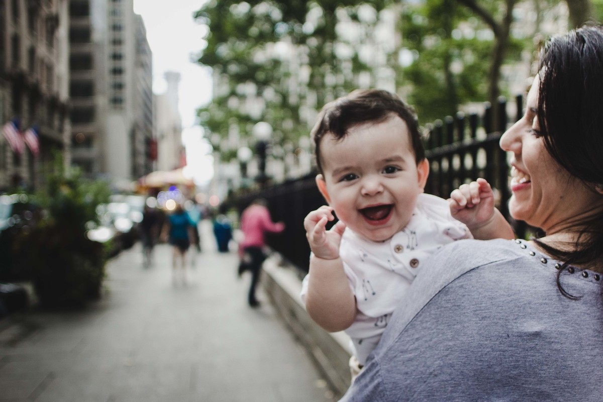 Ensaio fotográfico de família em Nova York, mãe segura filho no colo, criança olha para câmera e sorri, eles caminham por uma calçada em uma cidade