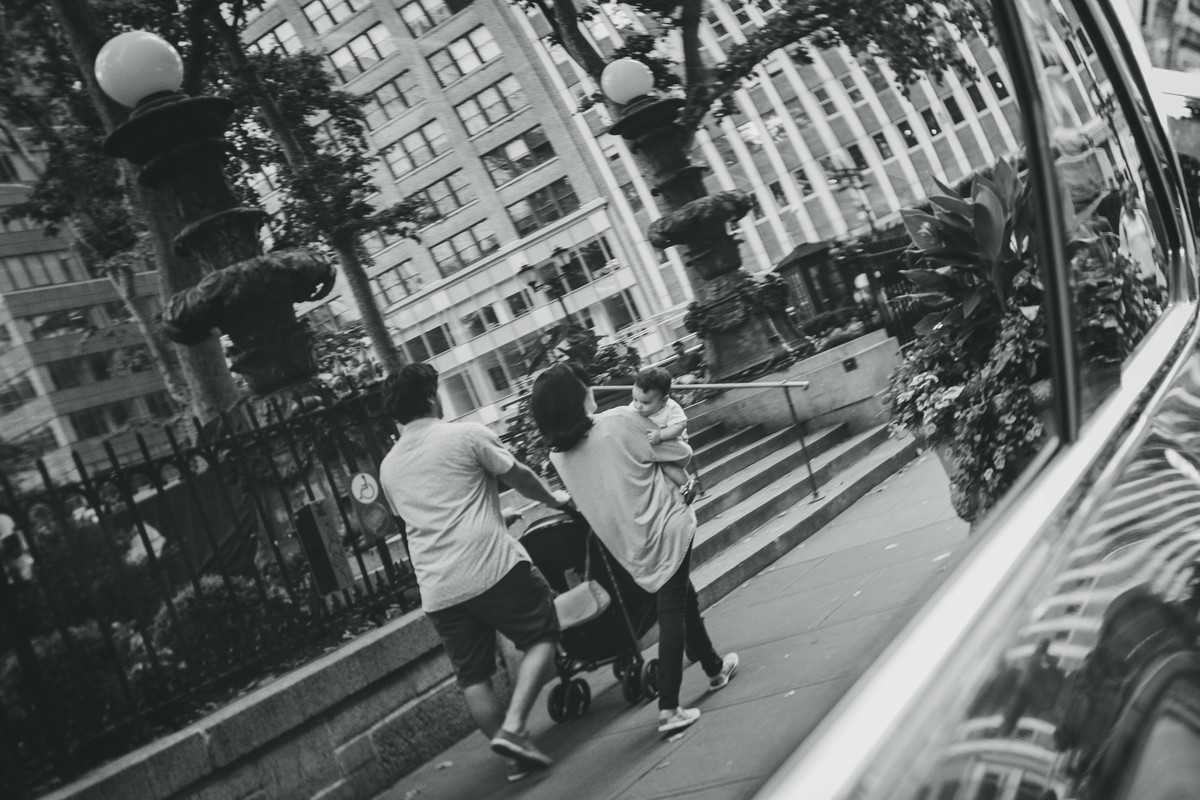 Ensaio fotográfico de família em Nova York, reflexo em vidro de um carro, mãe segura filho no colo, pai empurra carrinho de bebê, eles caminham por uma calçada em uma cidade