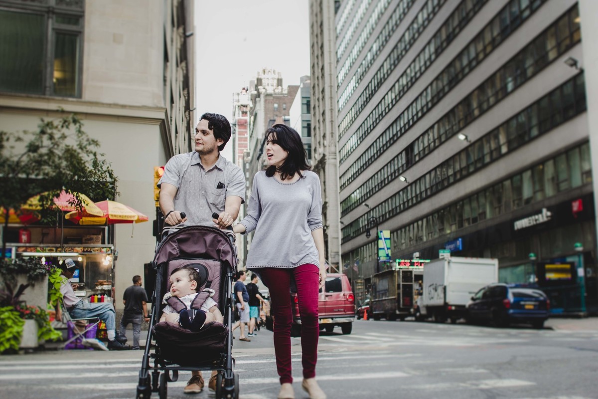 Ensaio fotográfico de família em Nova York, pai e mãe empurram carrinho de uma criança, eles atravessam a faixa de pedestres em uma grande cidade