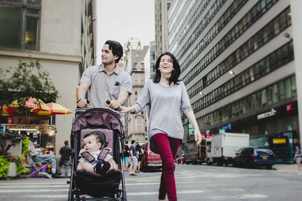 Ensaio fotográfico de família em Nova York, pai e mãe empurram carrinho de uma criança, eles atravessam a faixa de pedestres em uma grande cidade, mãe sorri