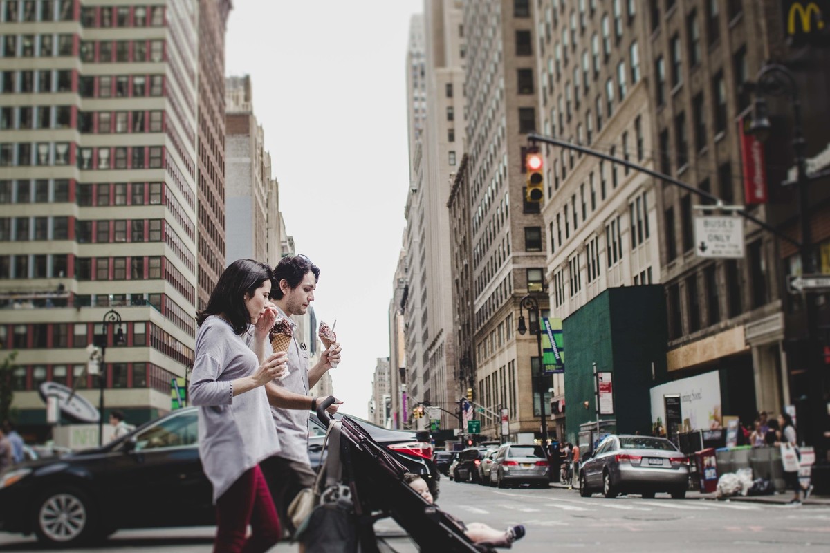 Ensaio fotográfico de família em Nova York, pai e mãe empurram carrinho de uma criança, eles atravessam a faixa de pedestres em uma grande cidade