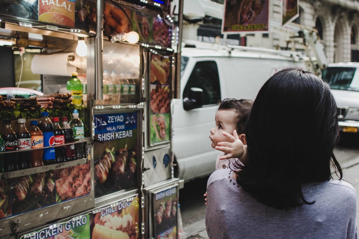 Ensaio fotográfico de família em Nova York, mãe segura filho no colo, criança olha para carrinho de cachoto quente, hot dog, eles caminham por uma calçada em uma cidade