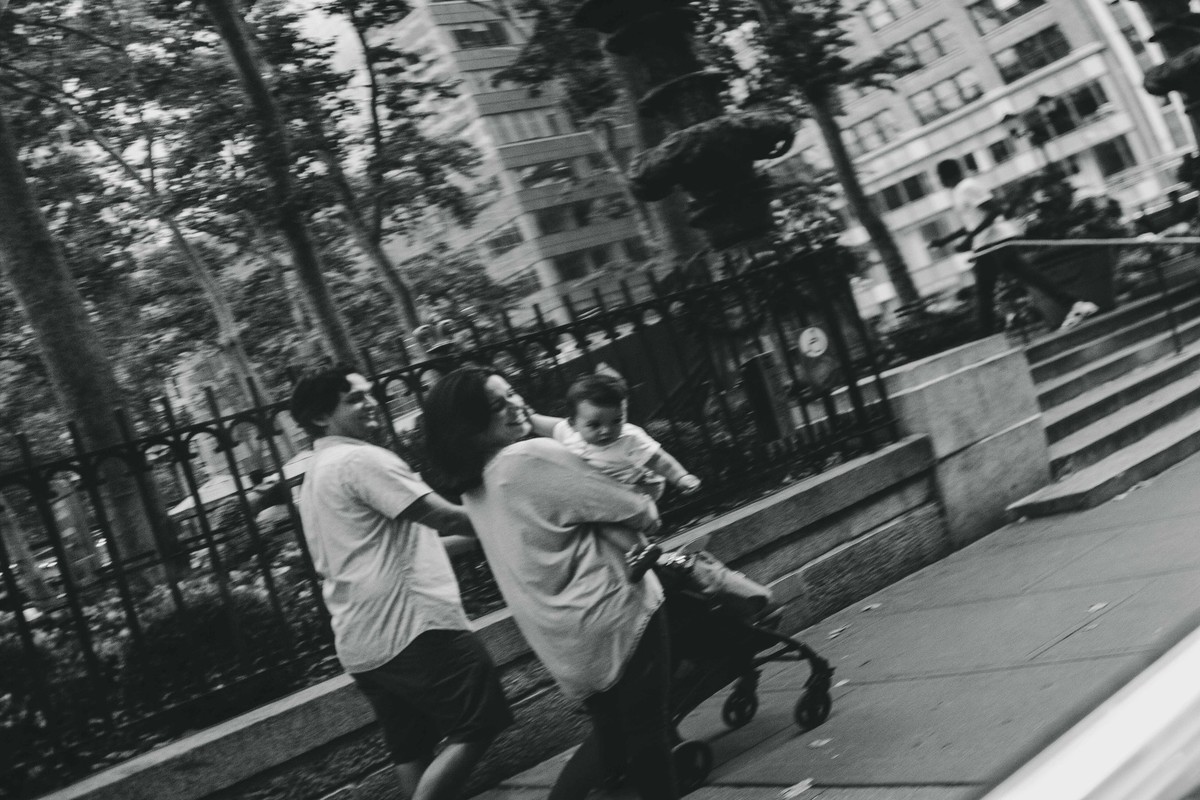Ensaio fotográfico de família em Nova York, reflexo em vidro de um carro, mãe segura filho no colo e olha para a câmera e sorri, pai empurra carrinho de bebê, eles caminham por uma calçada em uma cidade