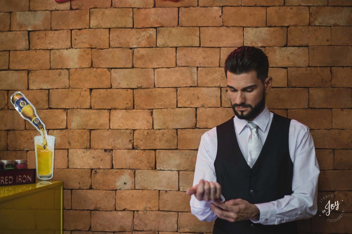 noivo dando os retoques finais para o casamento, ele está parado na frente de uma parede de tijolinhos e ajustando a blusa