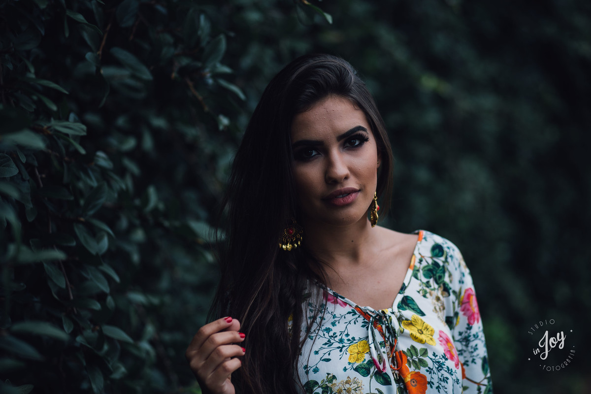 Fotografia feminina em que a modelo está segurando o cabelo e olhando para a camera na frente de um muro de cerca viva verde