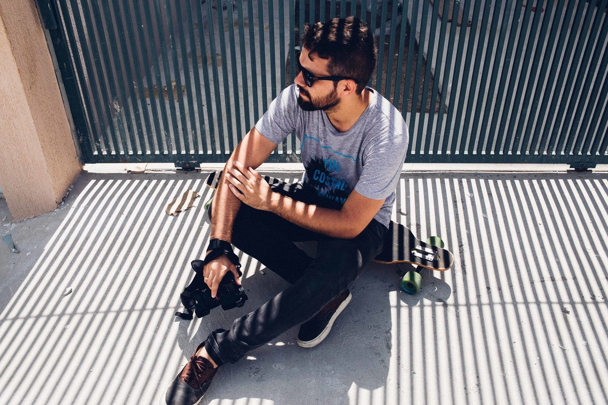 Ensaio masculino em que o modelo esta segurando uma camera e sentado encima de um longboard