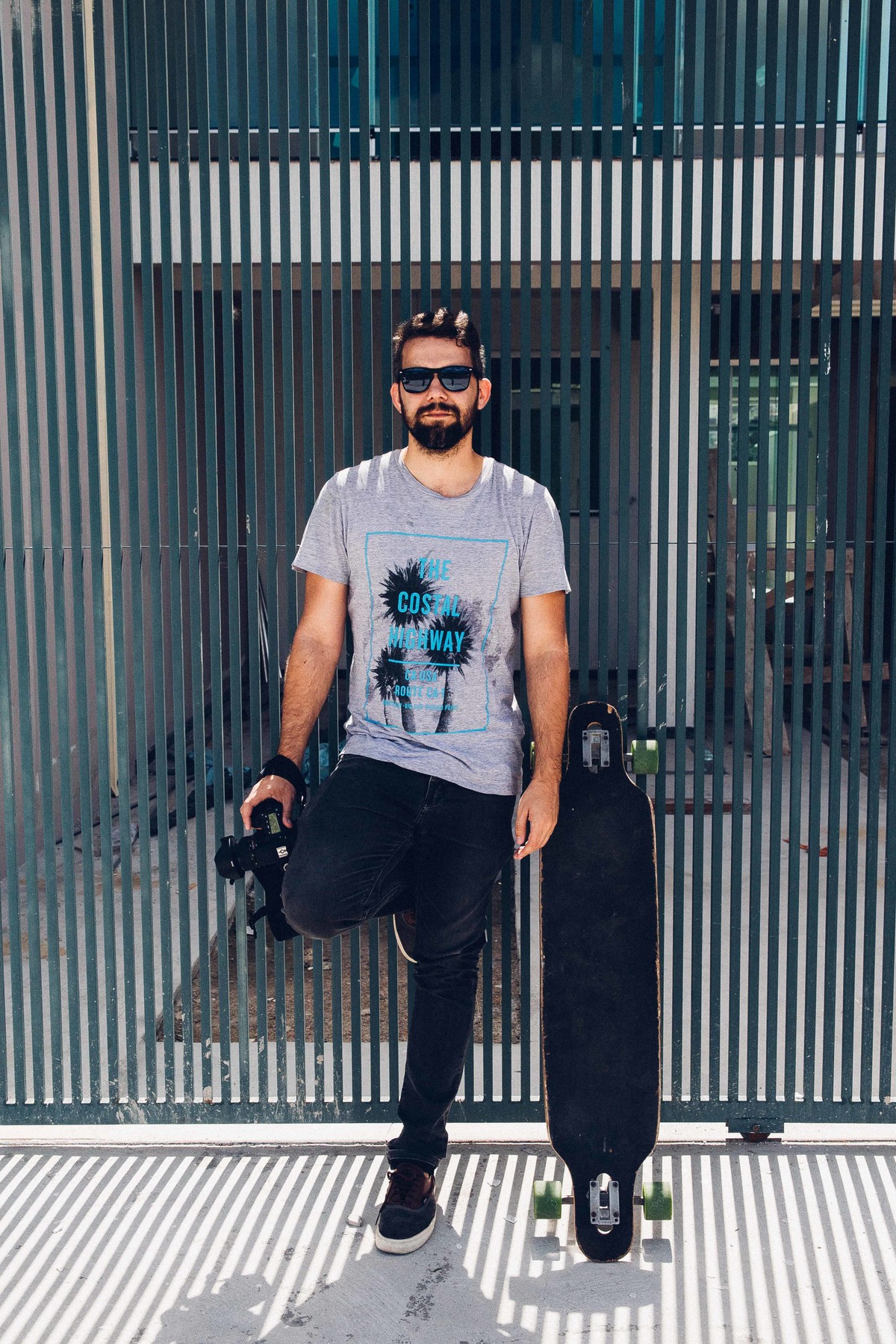 Ensaio masculino em que o modelo está de pé ao lado do longboard segurando uma camera