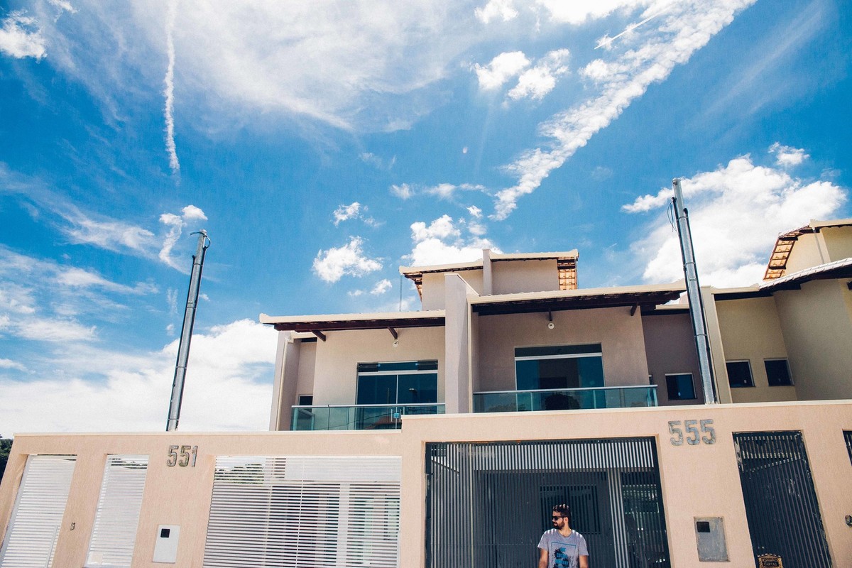 Ensaio masculino em que o modelo está de pé ao lado do longboard segurando uma camera