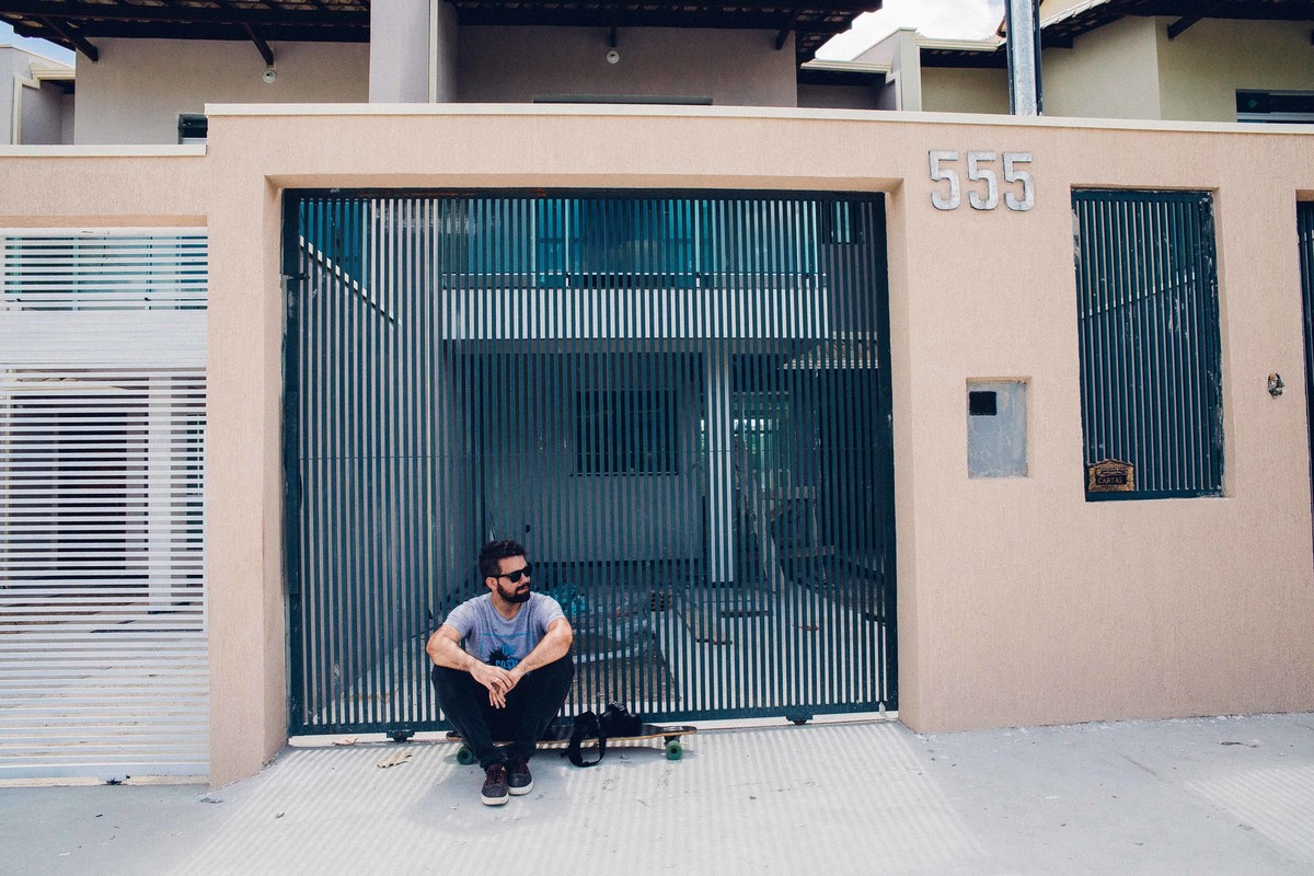 Ensaio masculino em que o modelo está de pé ao lado do longboard segurando uma camera