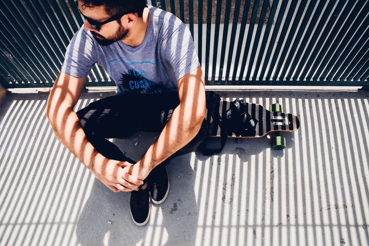 Ensaio masculino em que o modelo está de pé ao lado do longboard segurando uma camera