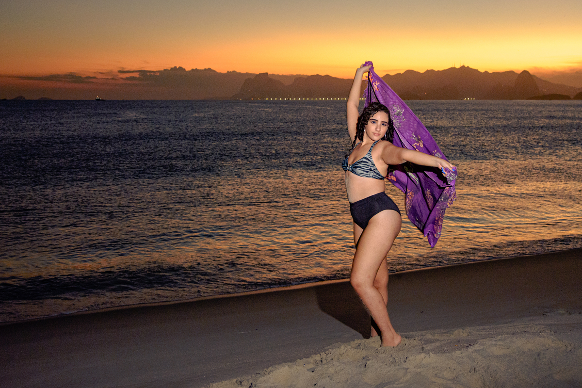 Fotografia da debutante ao por do sol com o mar ao fundo segurando canga, Rio de Janeiro - RJ