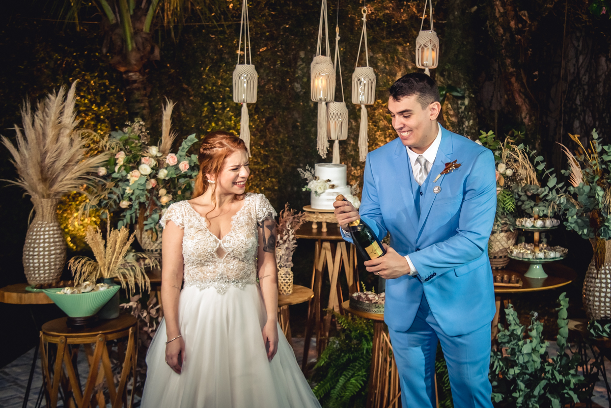 noivo abrindo a garrafa de Champagne com a noiva olhando e com a decoração da mesa do bolo no casamento no campo no sitio na barra da tijuca - RJ