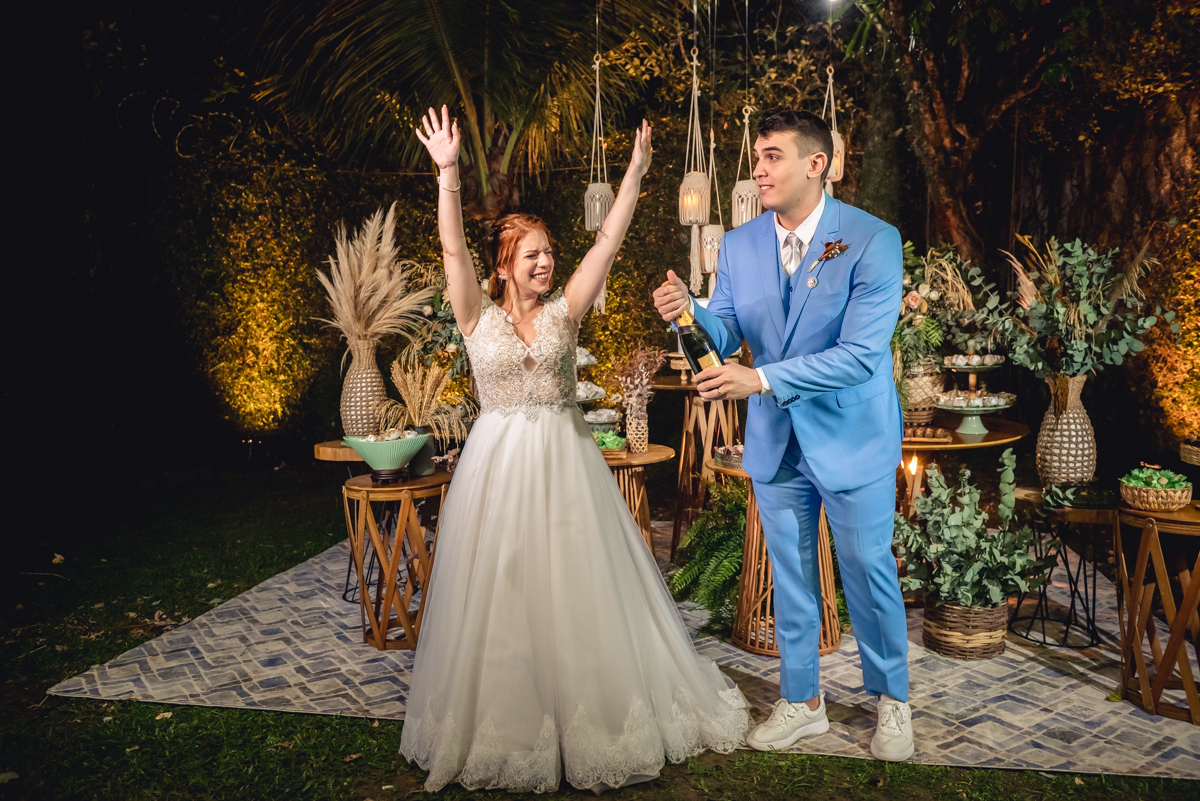 noiva com os braços abertos enquanto o noivo estoura o Champagne com a decoração da mesa do bolo no casamento no campo no sitio na barra da tijuca - RJ