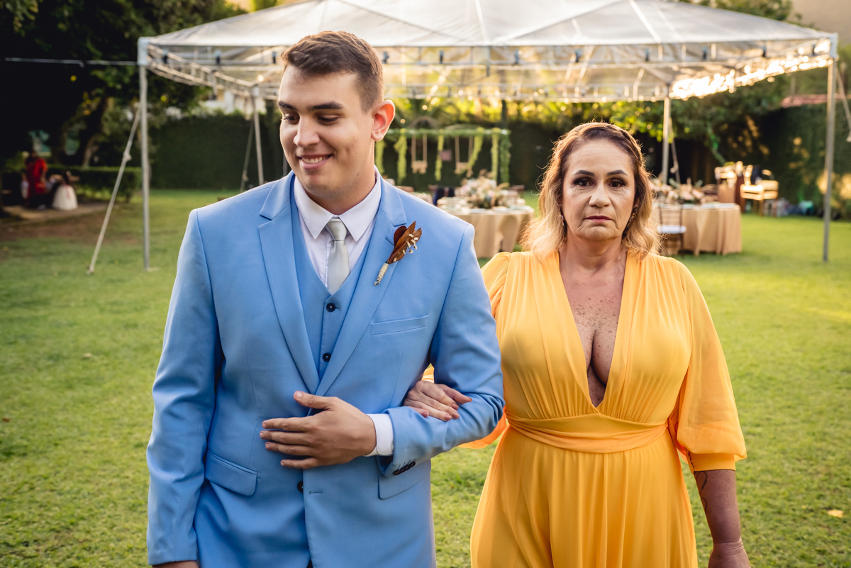 Entrada do noivo com terno azul e sua mãe com vestido amarelo no gramado verde para a cerimonia de casamento no sitio da paz, Barra da Tijuca - RJ