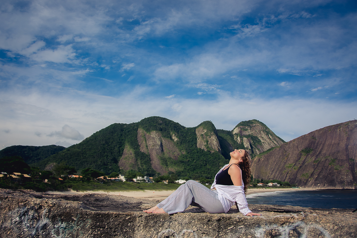 debutante sentada de perfil na pedra da praia de Itacoatiara com o céu azul de fundo rajado de nuvens