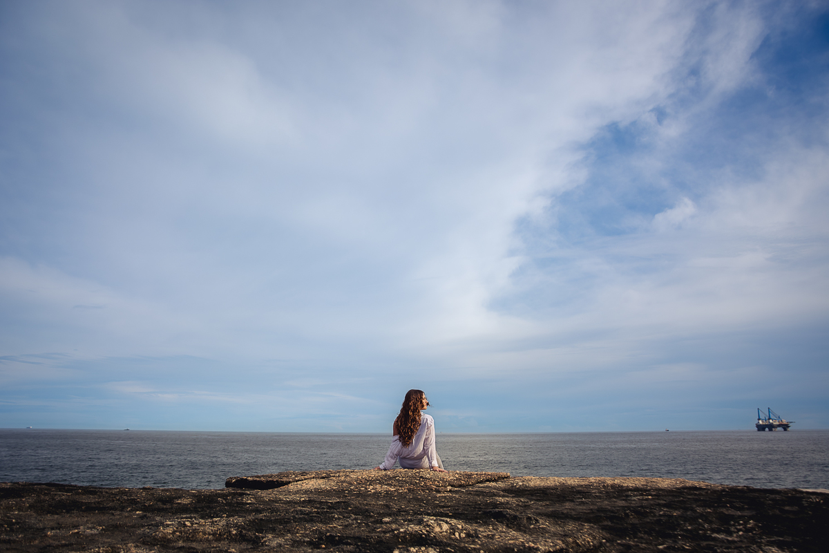 Debutante sentada na pedra de Itacoatiara, de frente para o mar, com o horizonte ao fundo e um belo céu azul, rajado de nuvens brancas
