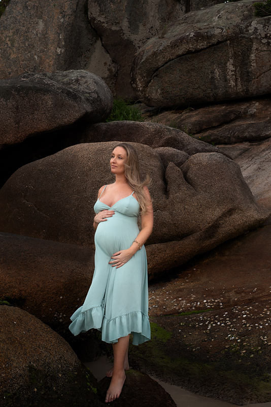 Gestante em pé nas pedras da Praia do Forte usando vestido verde água fotografada por Claudia Milani
