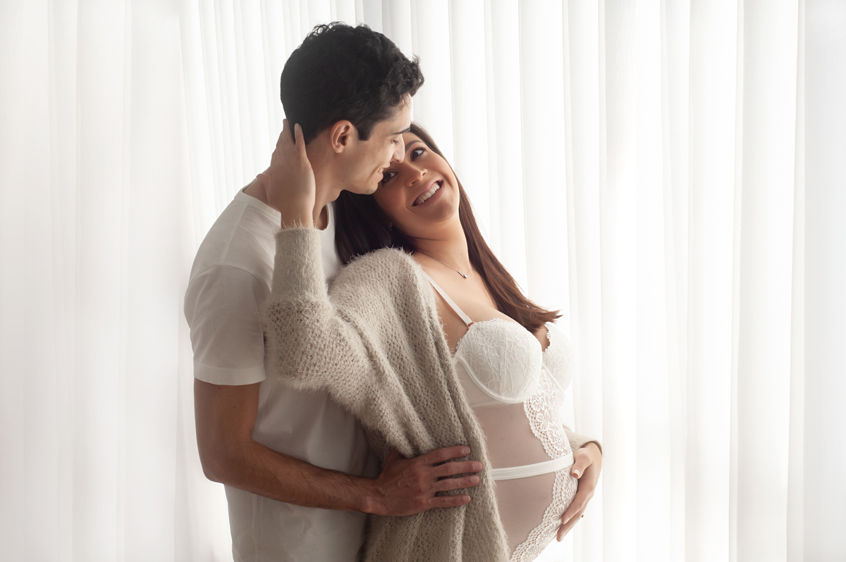Casal vestindo roupas claras ela está grávida ambos estão sorrindo enquanto ele envolve sua cintura e ela apoia a mão em seu rosto